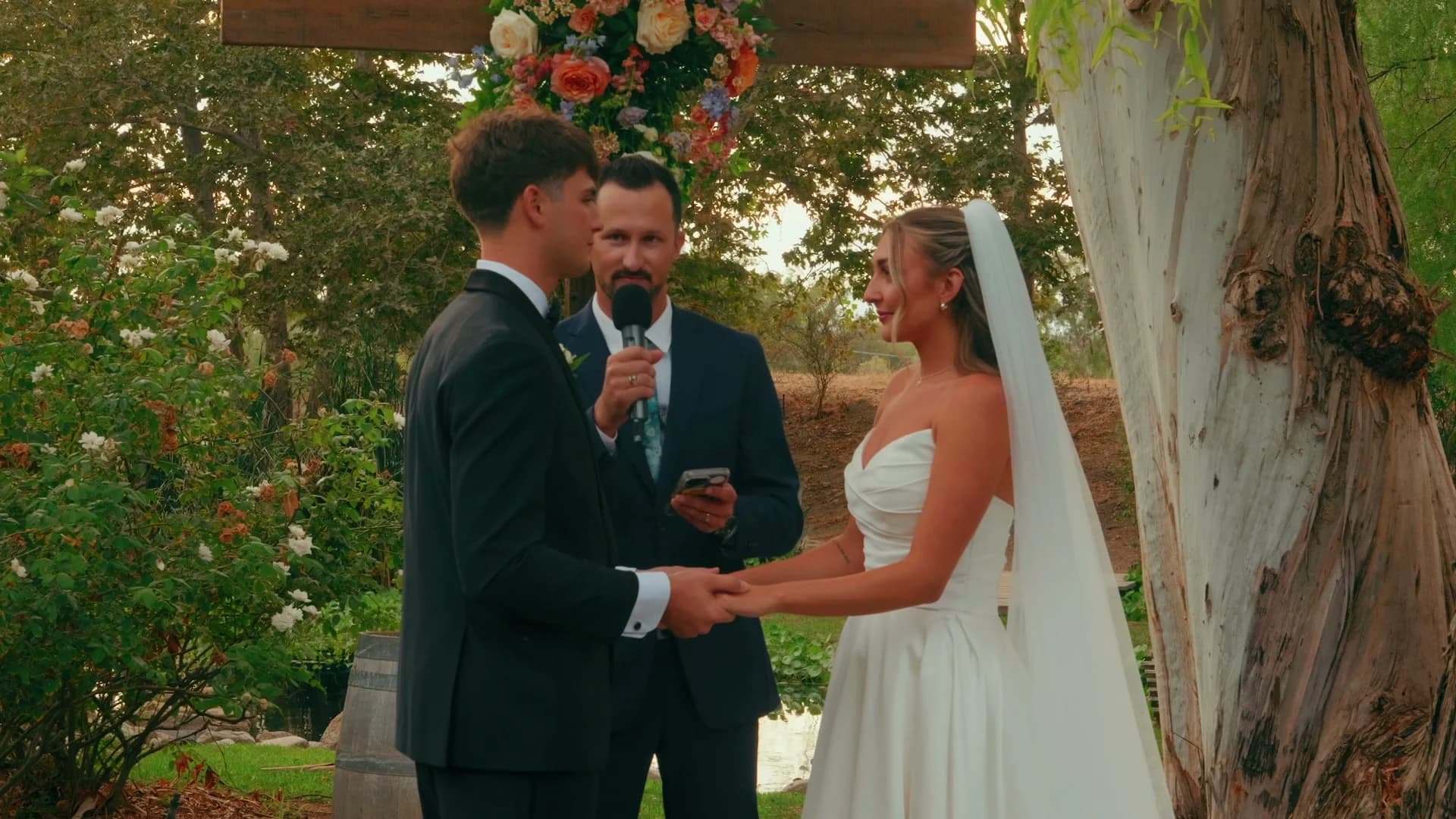 Denis + Cadin holding hands under the floral arch during the vows