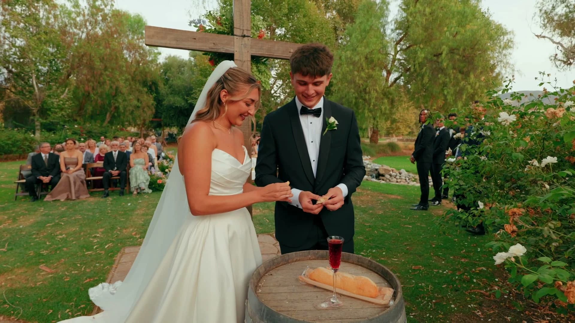 Denis + Cadin at a wine barrel with bread and wine, the wooden cross behind them