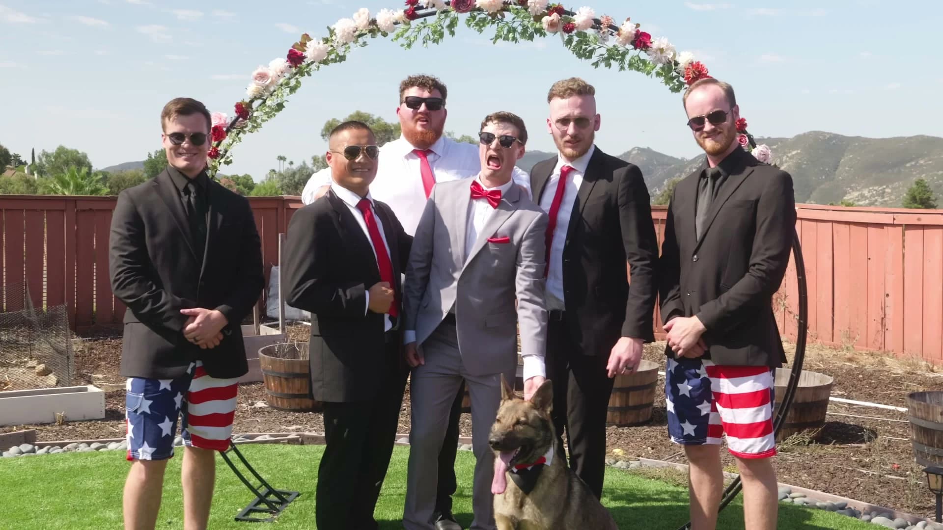 Groomsmen in flag shorts, and a dog, under the arch