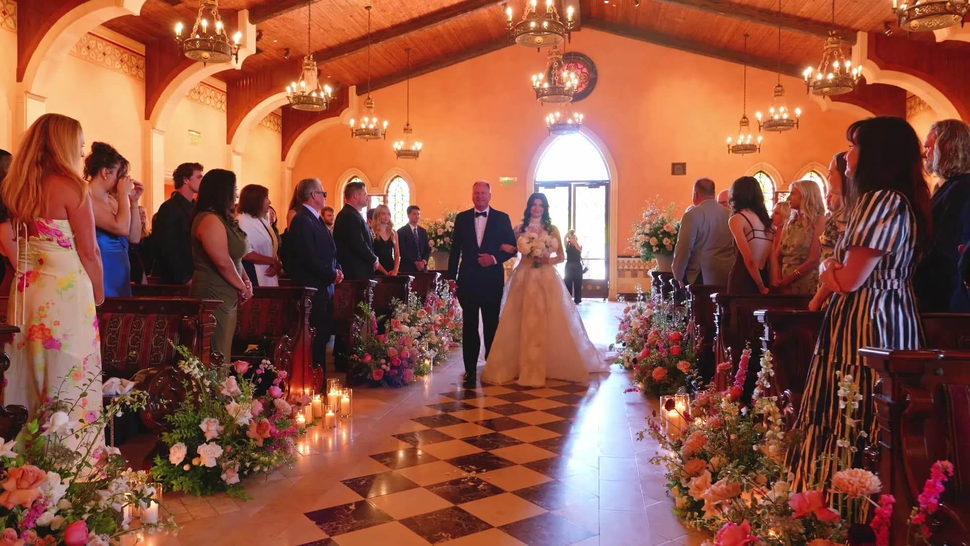 Kristen walking down the aisle past candles and pink flowers, chandeliers above