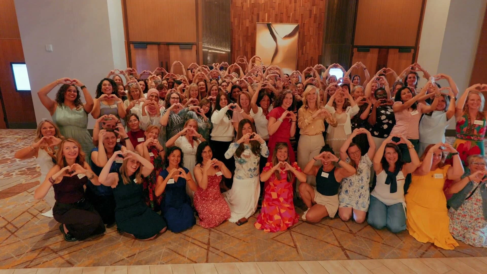 Several hundred conference attendees making heart shapes with their hands for a group photo in a hotel ballroom at Adored Wife Weekend, San Diego