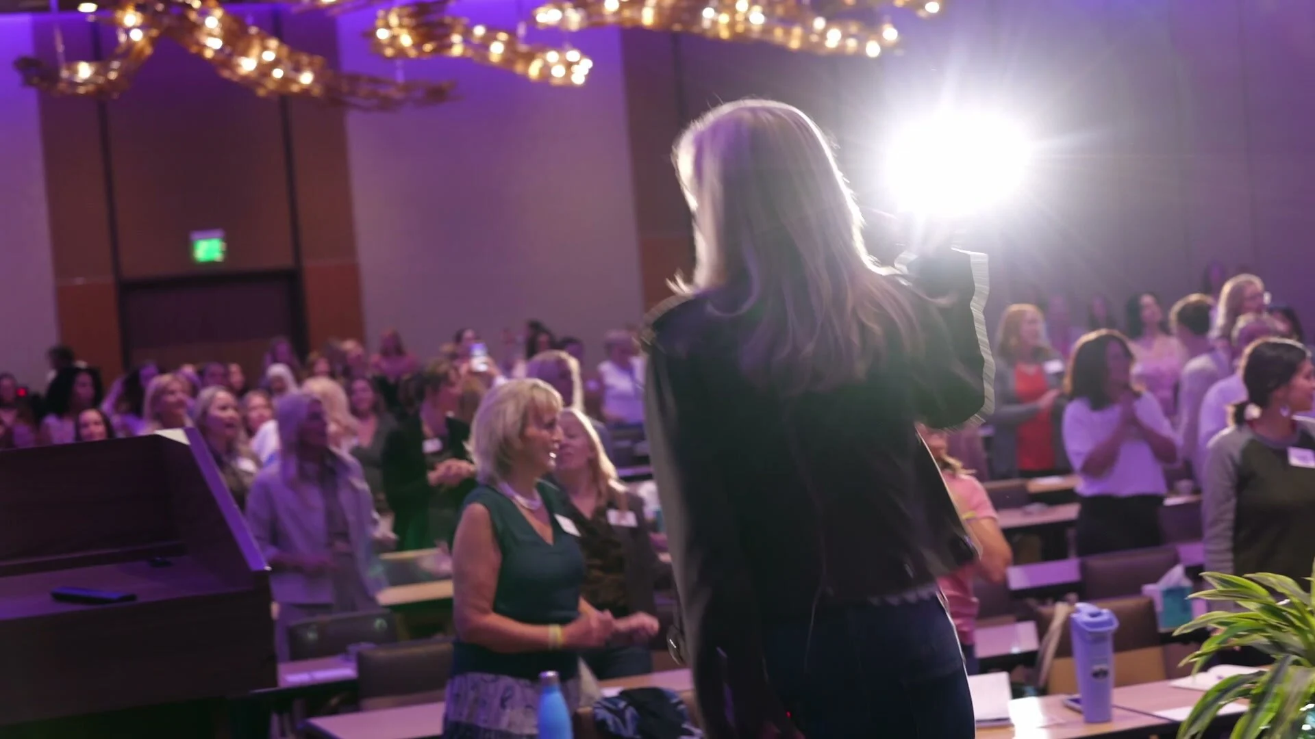 A speaker seen from behind, addressing a standing ballroom audience under stage lights at Adored Wife Weekend, San Diego