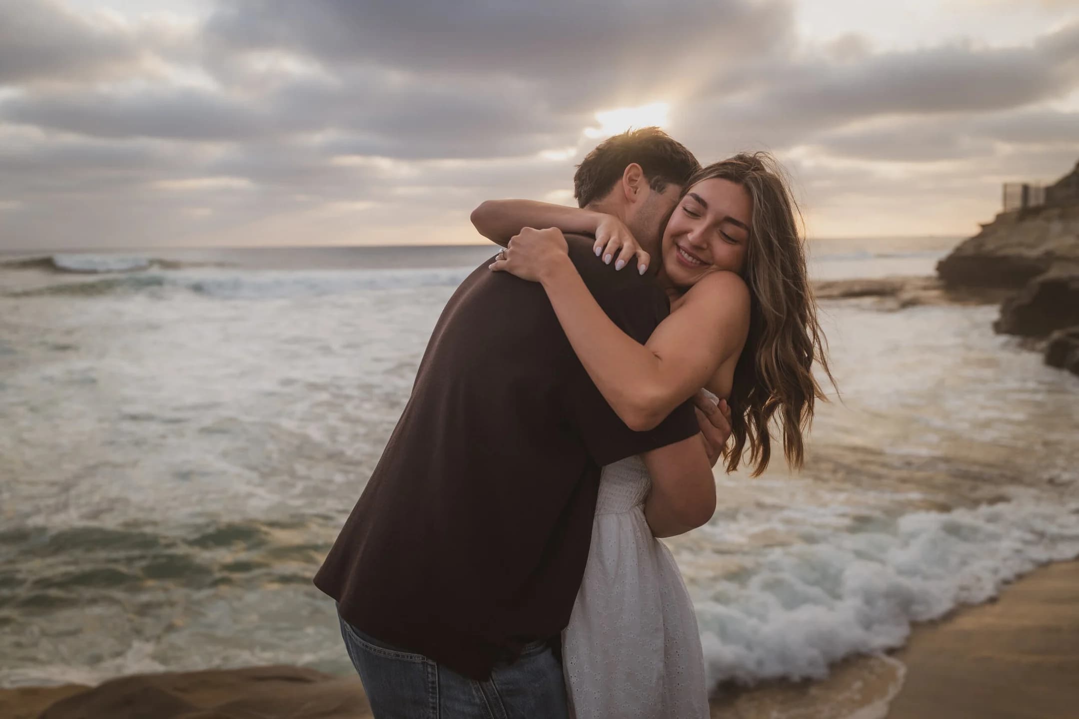 Denis + Cadin hug on the rocks by the ocean moments after the proposal — photographed by Paul Dal Studios