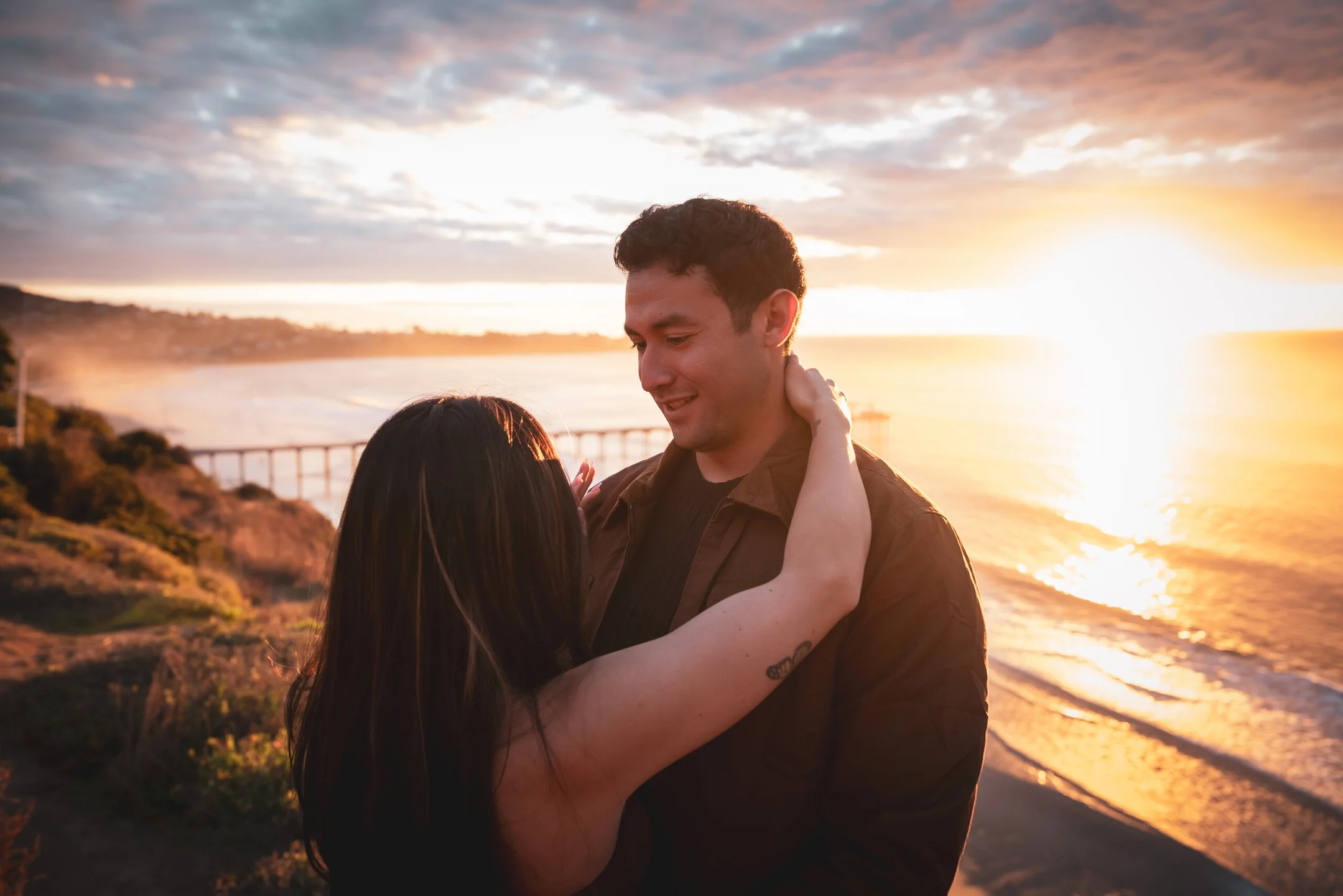 Mando + Melena embrace on the coastal bluffs above the pier at sunset after their proposal — photographed by Paul Dal Studios