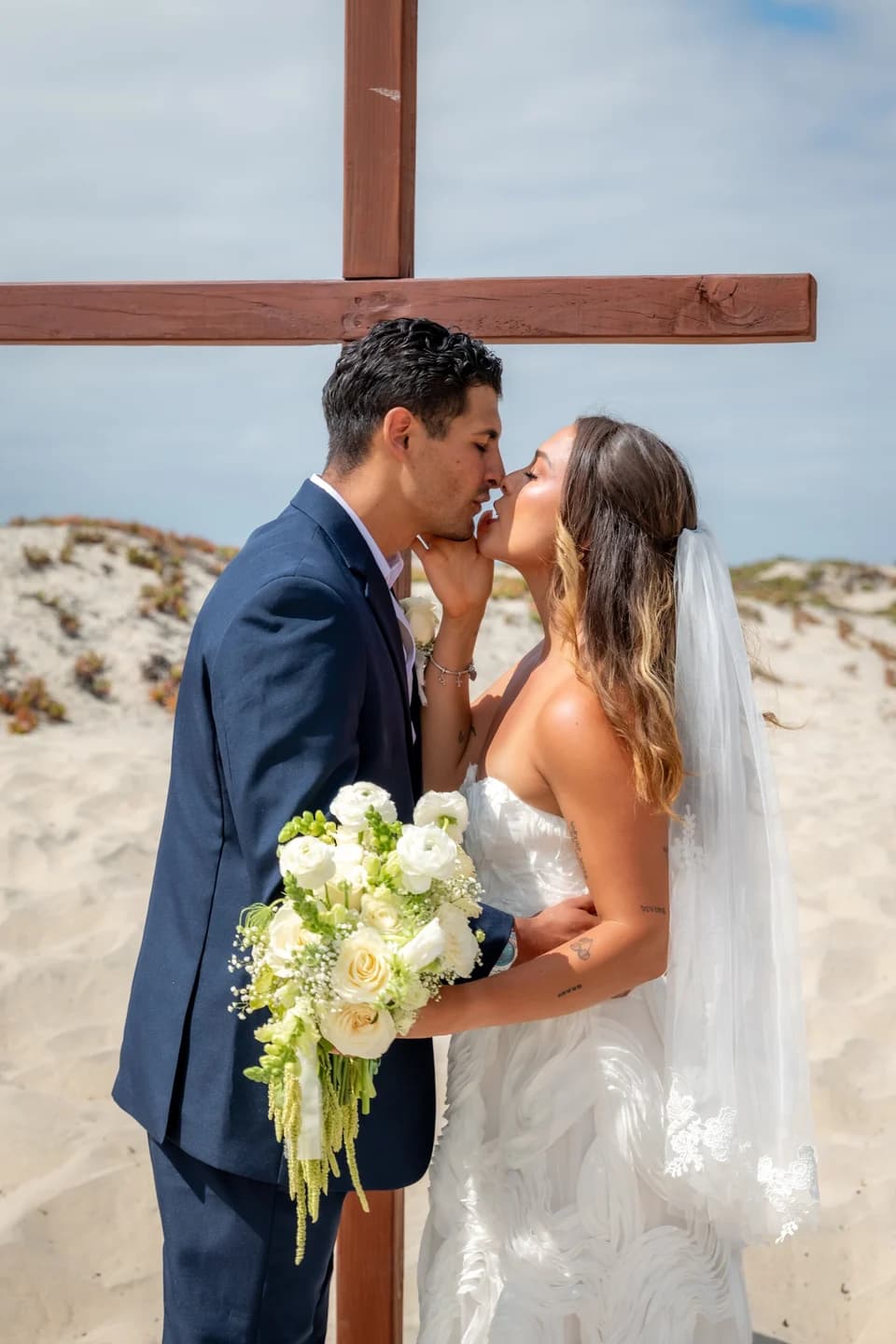 A bride and groom kiss beneath a wooden cross at their beach ceremony in San Diego — photographed by Paul Dal Studios